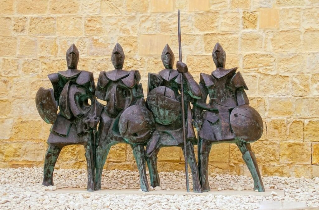 Modern bronze sculpture of four armoured knights with shields and spears, displayed against a sandstone wall in Malta