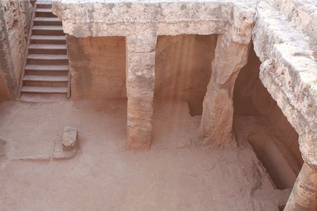 Sunlight filtering into a rock-cut burial chamber at the Tombs of the Kings in Paphos, Cyprus, with stone columns and stairs leading down.